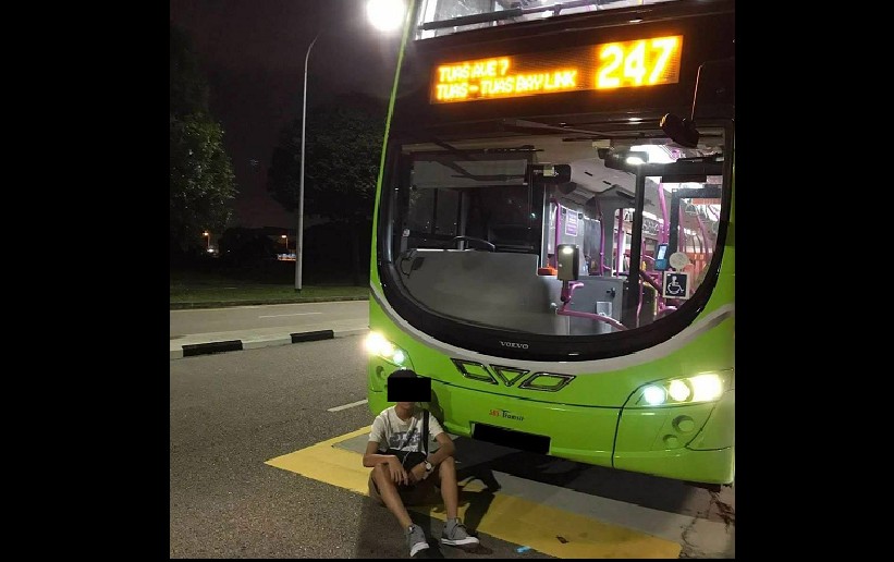 YOUTH POSES IN FRONT OF BUS, WHAT IS HE TRYING TO ACHIEVE?